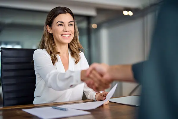 Business woman shakes hands with someone, papers in front of her on the desk