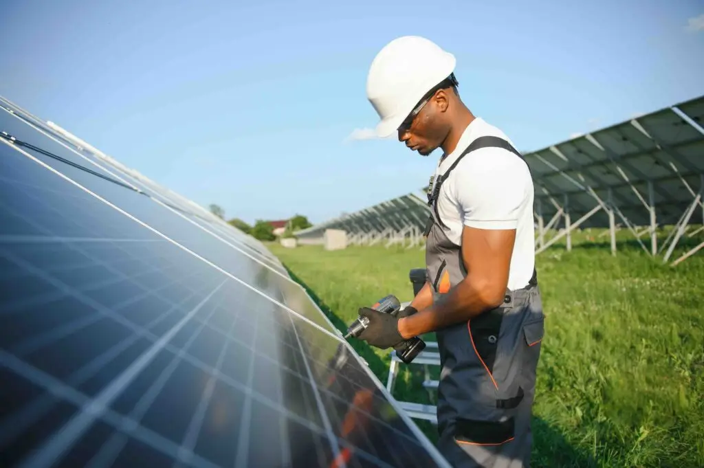 African american man in safety helmet and glasses tighten nuts on solar panels with screwdriver. Competent technician using tools while performing service work on station.