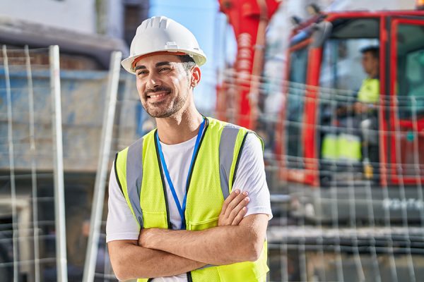 A young adult male in construction clothing smiles and crosses his arms